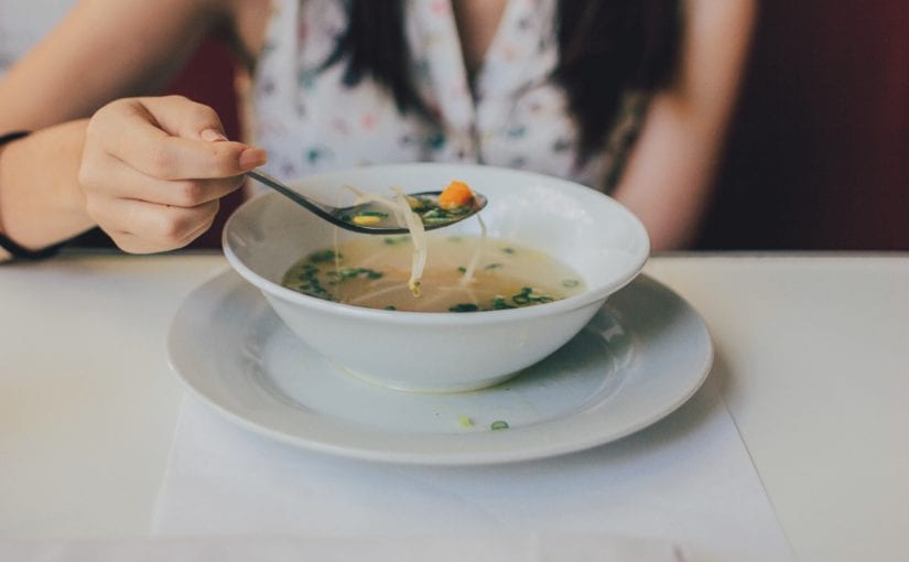 White glass bowl filled with soup on white glass plate on white table with women holding a spoon full of soup over bowl.