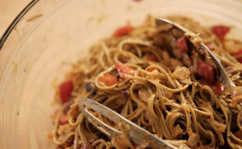 Clear glass bowl of pasta mixed with sauce and sliced tomatoes with mixing tongues.