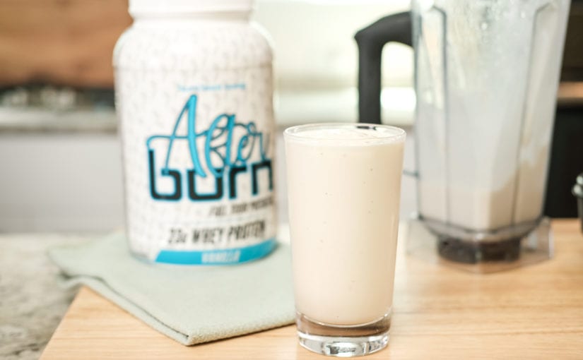 Clear glass filled with pina colada protein smoothie on cutting board on countertop next to white vanilla Afternoon bottle and empty blender.
